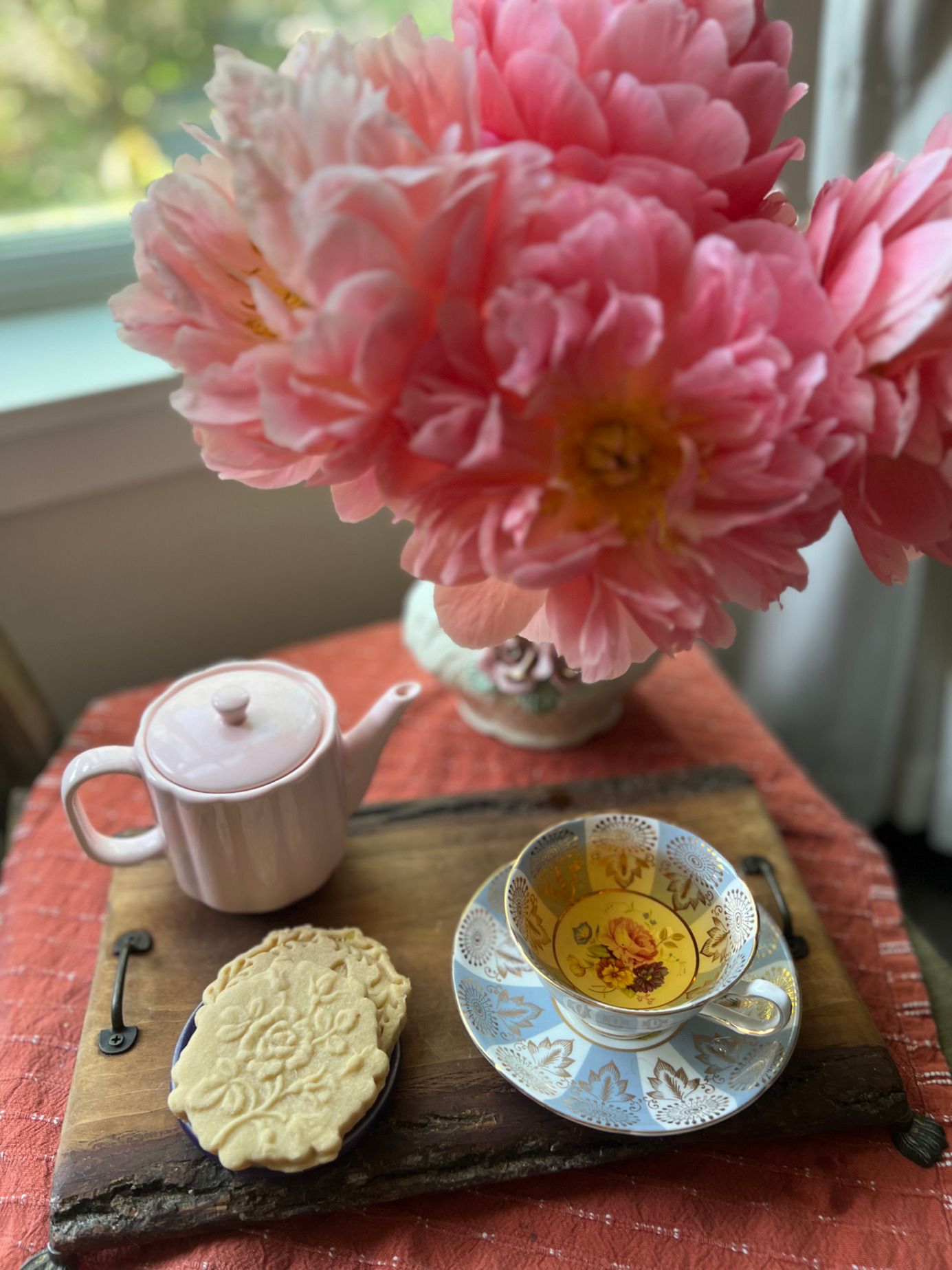 Peonies, a pink teapot, a cup of tea and a pressed shortbread cookie on a wooden tray.