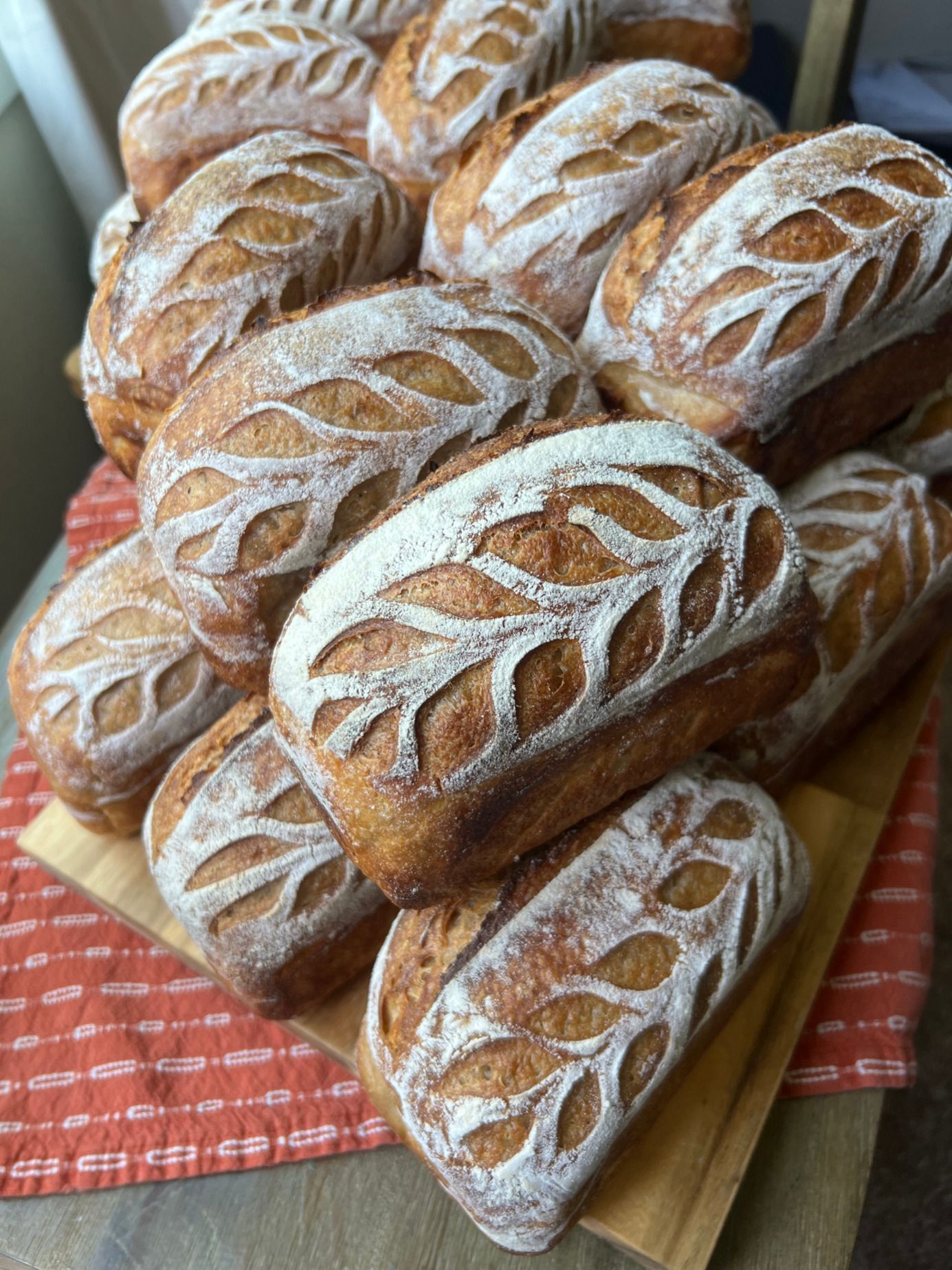 A pile of sourdough sandwich loaves, each scored with a stalk pattern and dusted with flour, on a red cloth.